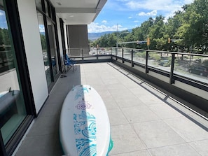 Un balcon avec une planche de surf, une rambarde en verre et une vue sur une route et des arbres.