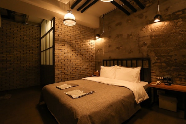Een kamer met een bakstenen muur, een bed met wit beddengoed, een nachtkastje met een lamp en een houten bankje.