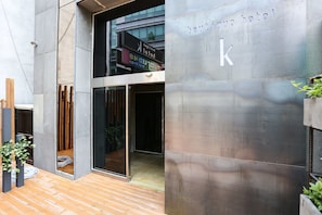 Een modern gebouw met een grote glazen ingang, een zichtbaar bord met 'boutique hotel k', en een houten terras.