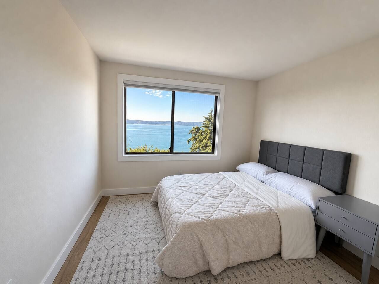 A bedroom with a large window, a bed with a grey headboard, and a bedside table.
