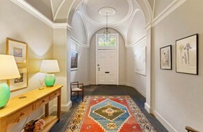 Un corridoio con tappeto decorato, un tavolino consolle in legno con lampade, opere d'arte incorniciate alle pareti e una porta bianca in fondo.