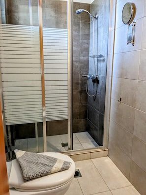 A bathroom with a toilet, a shower with a glass door, and a towel on the seat. - Villa Karina Near Achladies Beach (Skiathos)