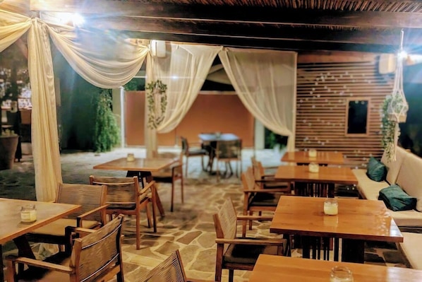 An outdoor dining area with wooden tables and chairs, draped white curtains, and a stone floor. - Villa Karina Near Achladies Beach (Skiathos)