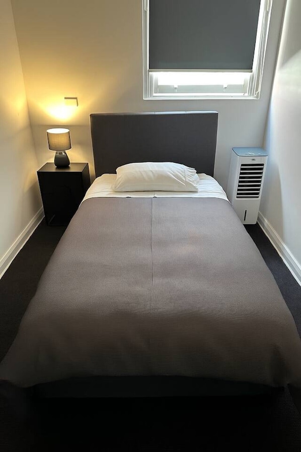 A single bed with a grey mattress and white bedding, a bedside table with a lamp, a window with blinds, and a wall-mounted air conditioning unit. - 5 mins to Central Sydney Lord Raglan Hotel (Alexandria)