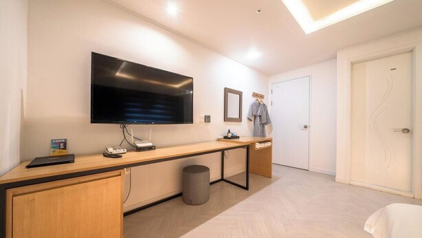 A modern hotel room with a large flat-screen TV mounted on the wall, a wooden desk with a laptop and a telephone, a small trash bin, and a door with a towel rack. - Suwon Hash Hotel (Suwon)
