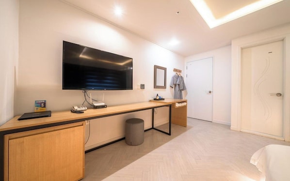 A modern hotel room with a large flat-screen TV mounted on the wall, a wooden desk with a laptop and a telephone, a small trash bin, and a door with a towel rack. - Suwon Hash Hotel (Suwon)