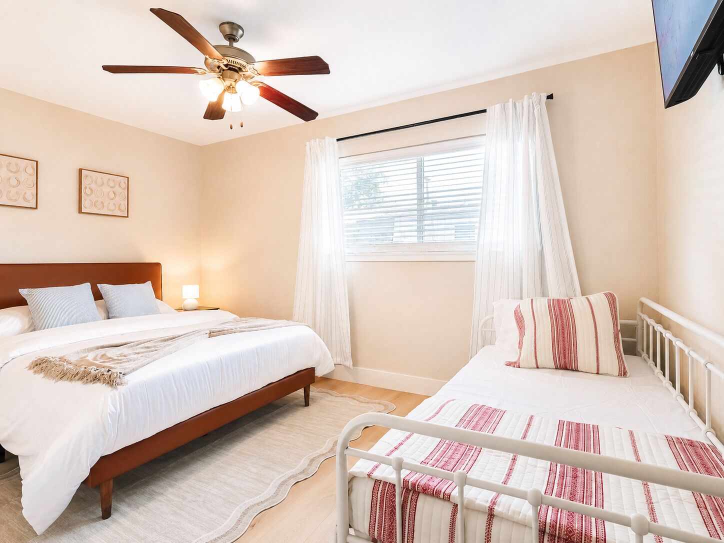 A bedroom with two beds, a ceiling fan, a window with curtains, and a TV mounted on the wall.