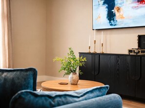 Ett modernt vardagsrum med ett runt soffbord i trä, en vas med gröna växter, ett svart skåp med guldfärgade detaljer och en stor, abstrakt tavla på väggen.