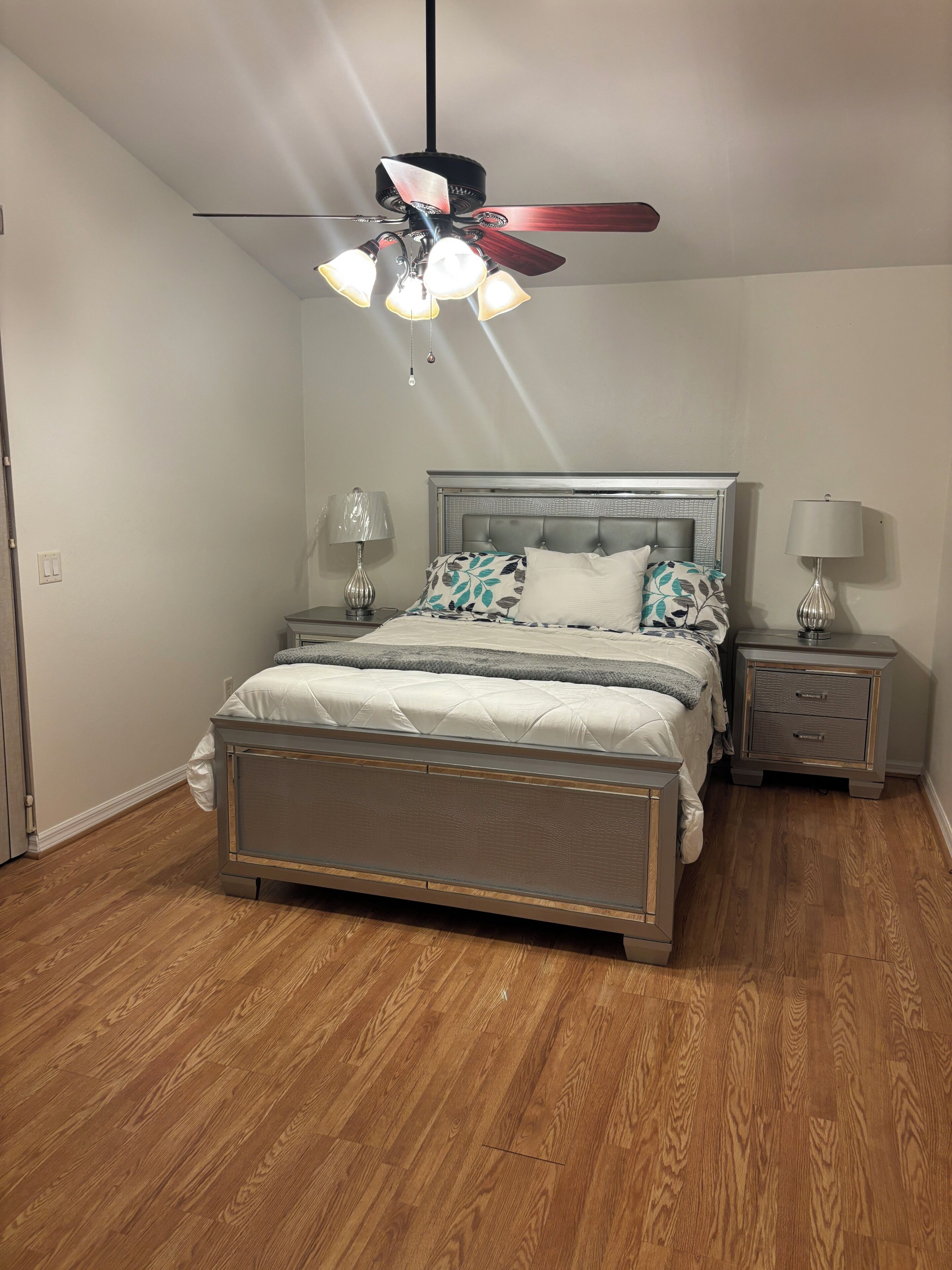 Un dormitorio con cama, mesitas de noche, ventilador de techo y suelo de madera.