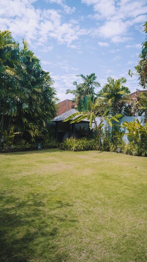 Un jardín bien cuidado con diversas plantas tropicales y una pequeña construcción, quizás un pabellón o cobertizo, al fondo.