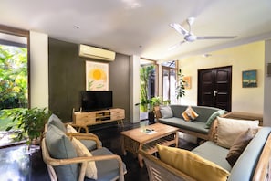 Ein Wohnzimmer mit Fernseher, einer Couch, Sesseln, einem Couchtisch und einem Deckenventilator.