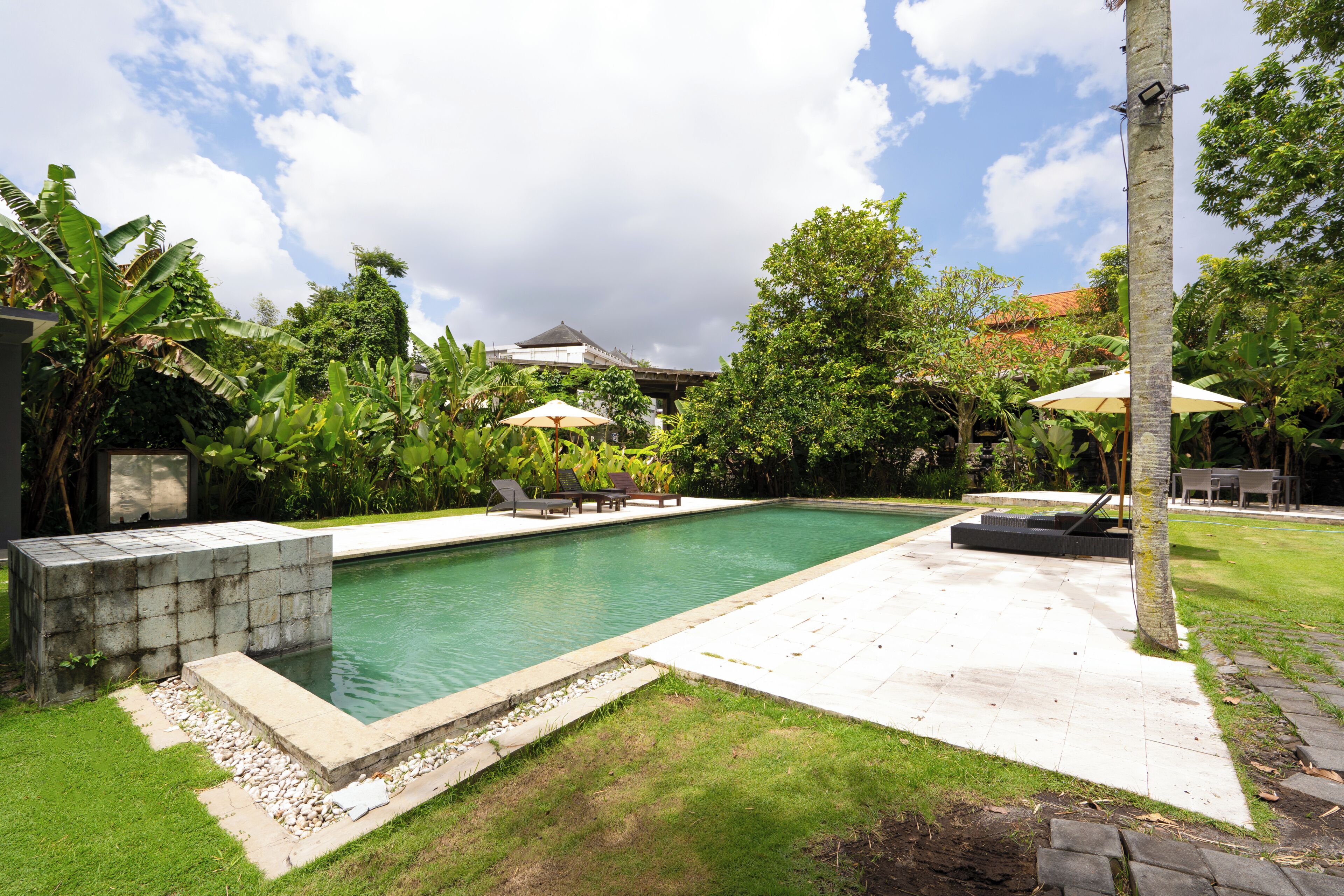 A swimming pool with lounge chairs and umbrellas, surrounded by lush greenery and a building in the background.