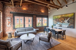 A spacious room with a brick wall, large windows, a gray sofa, a round coffee table, and a dining area with chairs and a table. - Grand Loft Collection | Up to 19 Guests (Asheville)