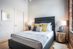 Een moderne slaapkamer met een groot bed, twee nachtkastjes, een wandlamp, een ingelijst kunstwerk en een bakstenen muur.