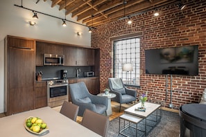 A modern kitchen with built-in appliances, a dining area with a table and chairs, and a living space with a sofa and a TV mounted on a brick wall. - Grand Loft Collection | Up to 19 Guests (Asheville)