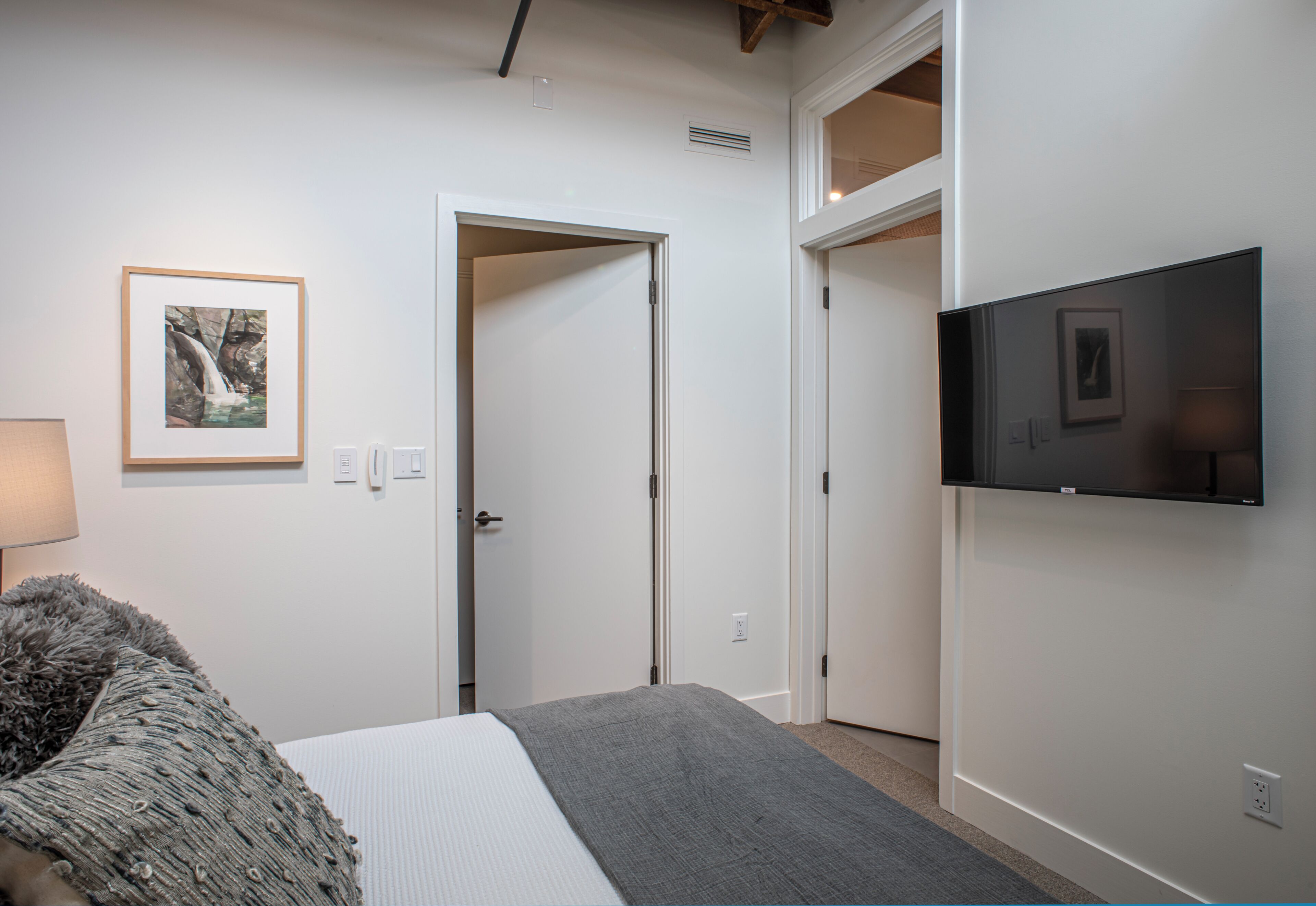 Un dormitorio con cama, televisor de pantalla plana montado en la pared, puerta que da a otra habitación y un cuadro enmarcado.
