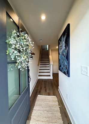 Un pasillo con piso de madera, pared blanca, un cuadro abstracto negro y una escalera que conduce a otro nivel.