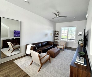 A modern living room with a leather sofa, a wooden coffee table, a side table with a book, a floor lamp, and a large mirror. - Modern Townhome - 2 Bedroom / 2 Bathroom - Beltline, Grant Park, Downtown, Zoo (Atlanta)