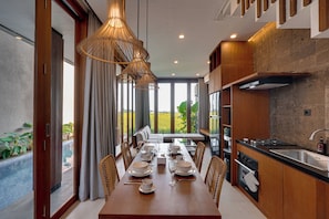 A modern kitchen with a dining area, large windows, and a view of the outdoors. - Villa Casa De Leonel - Kedungu (Tabanan)