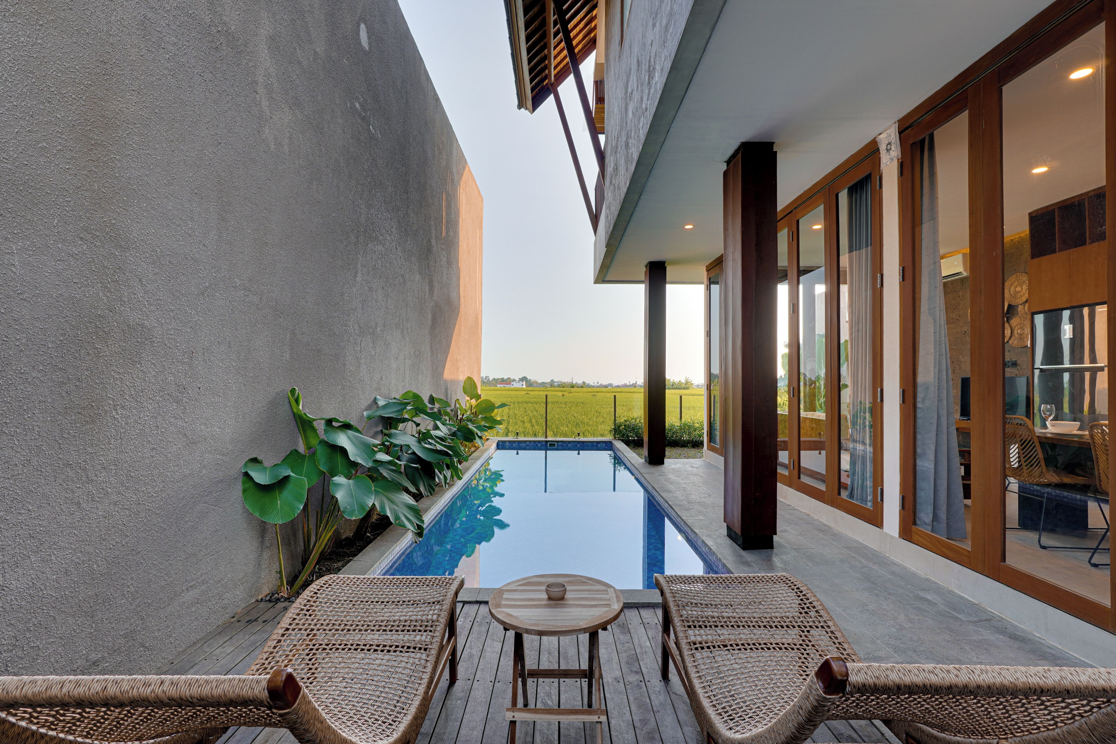 Ein moderner Poolbereich im Freien mit Blick auf ein grünes Feld, eine Holzterrasse mit zwei Liegestühlen und ein verglaster Essbereich.