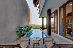 Un área de piscina moderna al aire libre con vista a un campo verde, una terraza de madera con dos sillones de descanso y un comedor con paredes de vidrio.