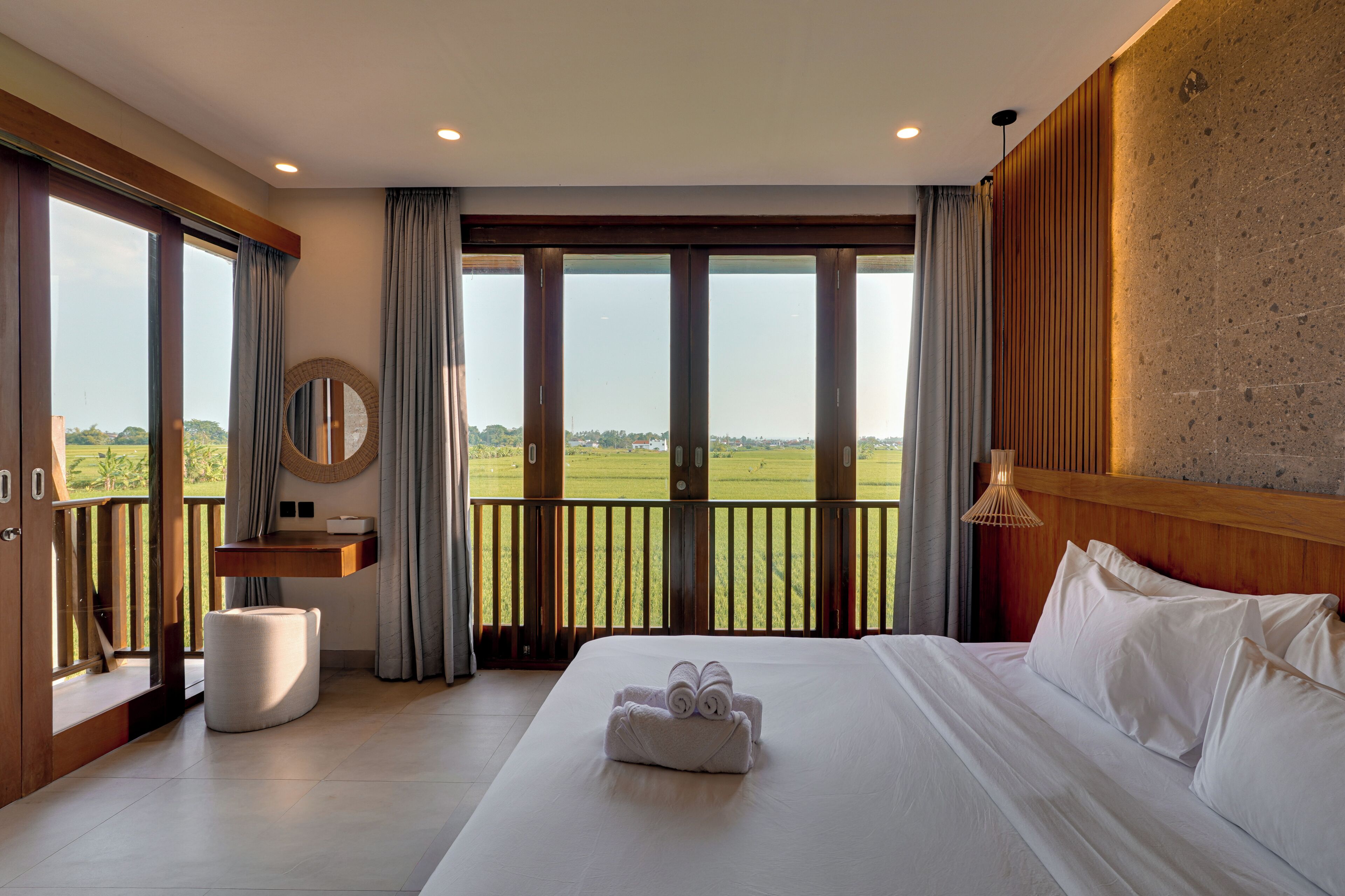 Habitación de hotel con una cama grande, cabecera de madera, espejo redondo, una mesita pequeña y vistas a un paisaje verde a través de ventanas amplias.