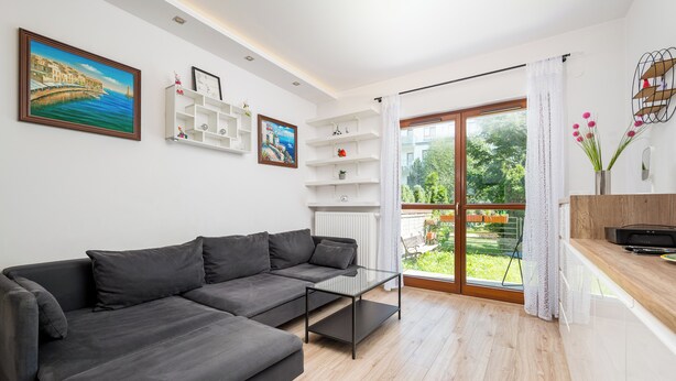 A modern living room with a grey sectional sofa, white shelving units, and a large window with a view of greenery. - Royal Wilanów Charming Apartment (Warszawa)