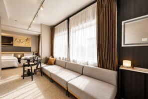 A hotel room with a sofa, a dining table, two beds, and a large window with curtains.