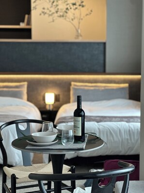 A hotel room with a dining table set for two, a bottle of wine, and a glass. The room has a bed with white bedding and a nightstand with a lamp.