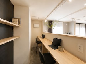 A modern office space with a long wooden desk, two black chairs, a laptop, and a mug. There are shelves with books and a plant on the windowsill. - KATAWARE  About 10 minutes to Shinjuku | 3-minute walk from Numabukuro Station | Private 1LDK | 60 ㎡ | Accommodates up to 6 people | 3-minute walk to a supermarket and convenience store (中野区)