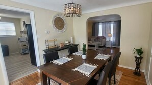 A dining area with a wooden table, chairs, and a chandelier. There is a kitchen area with a microwave and a trash bin.