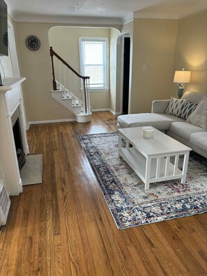Un salon doté d’un parquet, d’une table basse blanche, d’un canapé, d’un tapis à motifs, d’un escalier et d’une cheminée.