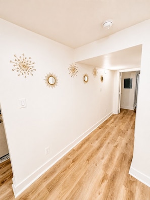 A hallway with white walls and wooden flooring, featuring decorative wall sconces and a ceiling light. - The Ascended Retreat (Dallas)
