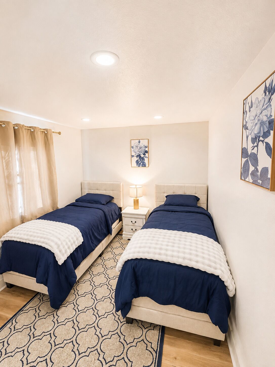 Une chambre avec deux lits, une table de chevet, une lampe et une œuvre d’art à motifs floraux accrochée au mur.