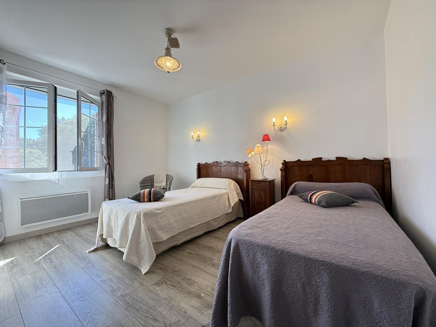 A hotel room with two beds, a window with curtains, a radiator, and a wall-mounted lamp.