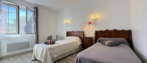 A hotel room with two beds, a window with curtains, a radiator, and a wall-mounted lamp.