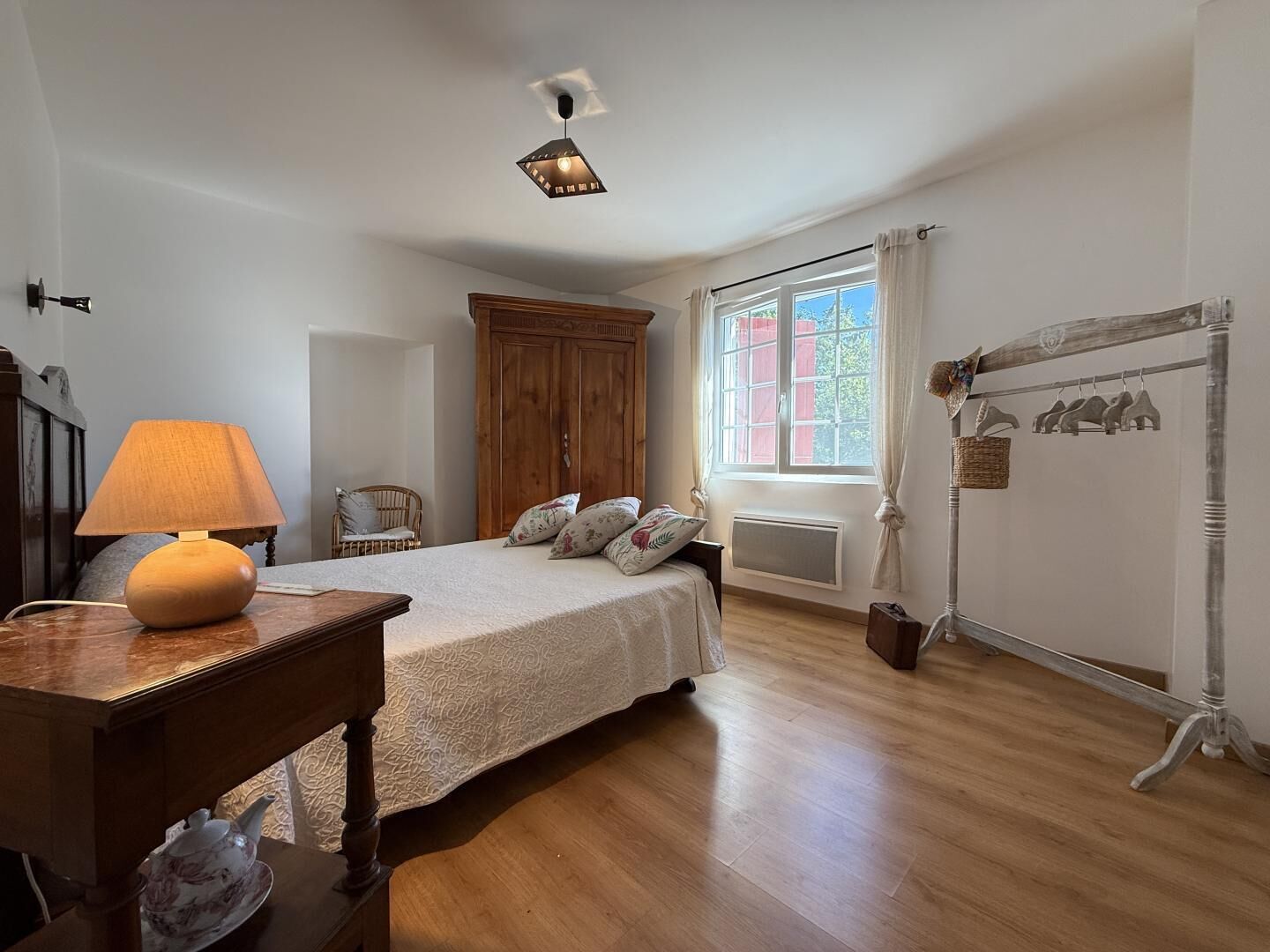 Un dormitorio con cama, una mesita de noche de madera, una lámpara, un armario, un radiador y una ventana con cortinas.