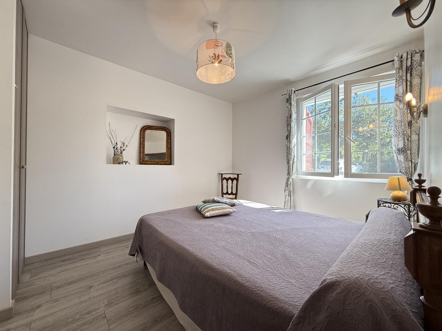 Un dormitorio con cama, espejo, ventana con cortinas y una lámpara colgante.