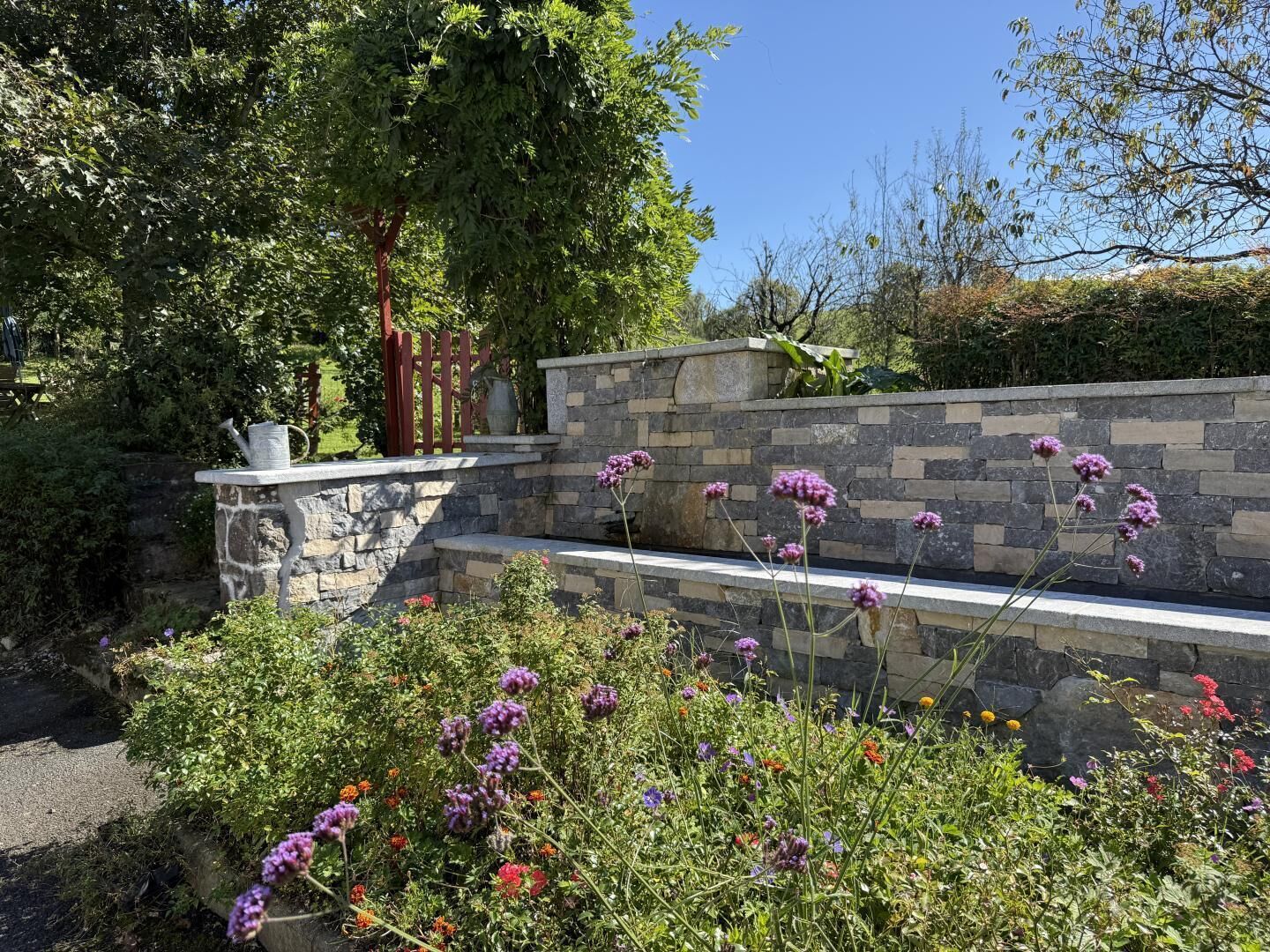 A garden with a stone wall, a metal gate, and various flowering plants.