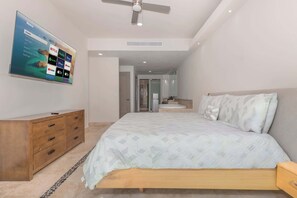 Quarto com cama, cômoda, ventilador de teto e TV fixada na parede exibindo serviços de streaming.