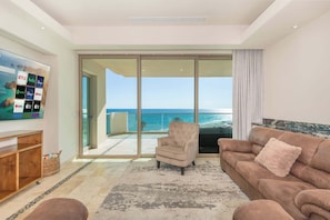 A living room with a television displaying streaming services, a sofa, a chair, and a view of the ocean through large glass doors. - Tessoro 204 Beachfront 3BR Luxury Escape (Las Conchas)