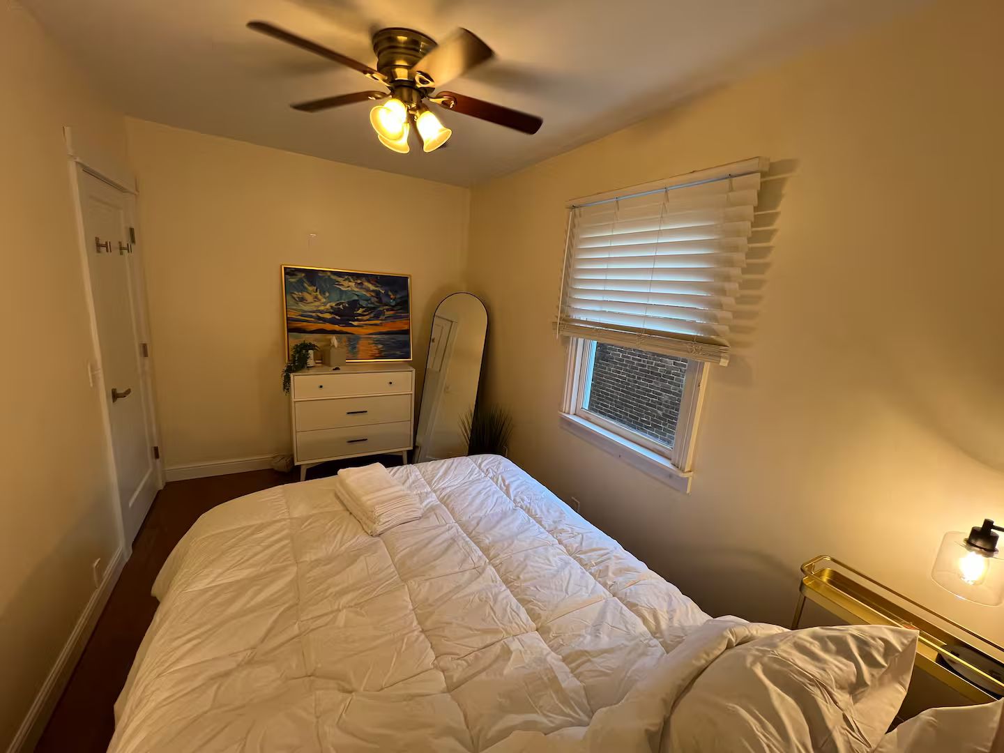 A bedroom with a bed, a ceiling fan, a dresser, a painting, a window with blinds, and a nightstand with a lamp.