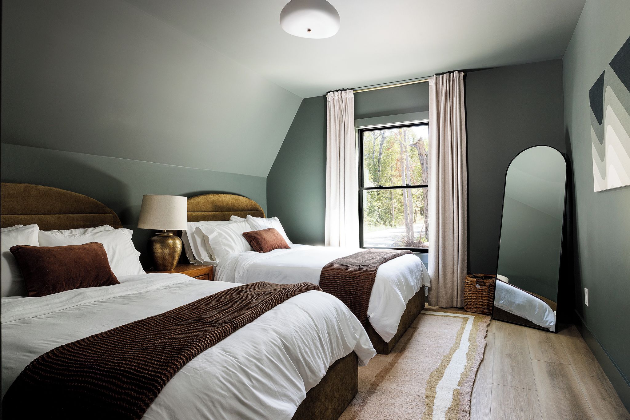 A bedroom with two beds, a nightstand, a lamp, a window with curtains, and a mirror.
