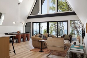 A spacious loft with a dining area, seating, and large windows offering a view of the outdoors. - Northport Noir | Waterfront Aframe Retreat (Northport)
