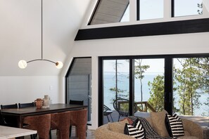 A modern interior with a dining area, seating by a large window overlooking a body of water. - Northport Noir | Waterfront Aframe Retreat (Northport)