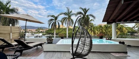 A rooftop pool area with lounge chairs, a hanging chair, and a view of palm trees and a marina.