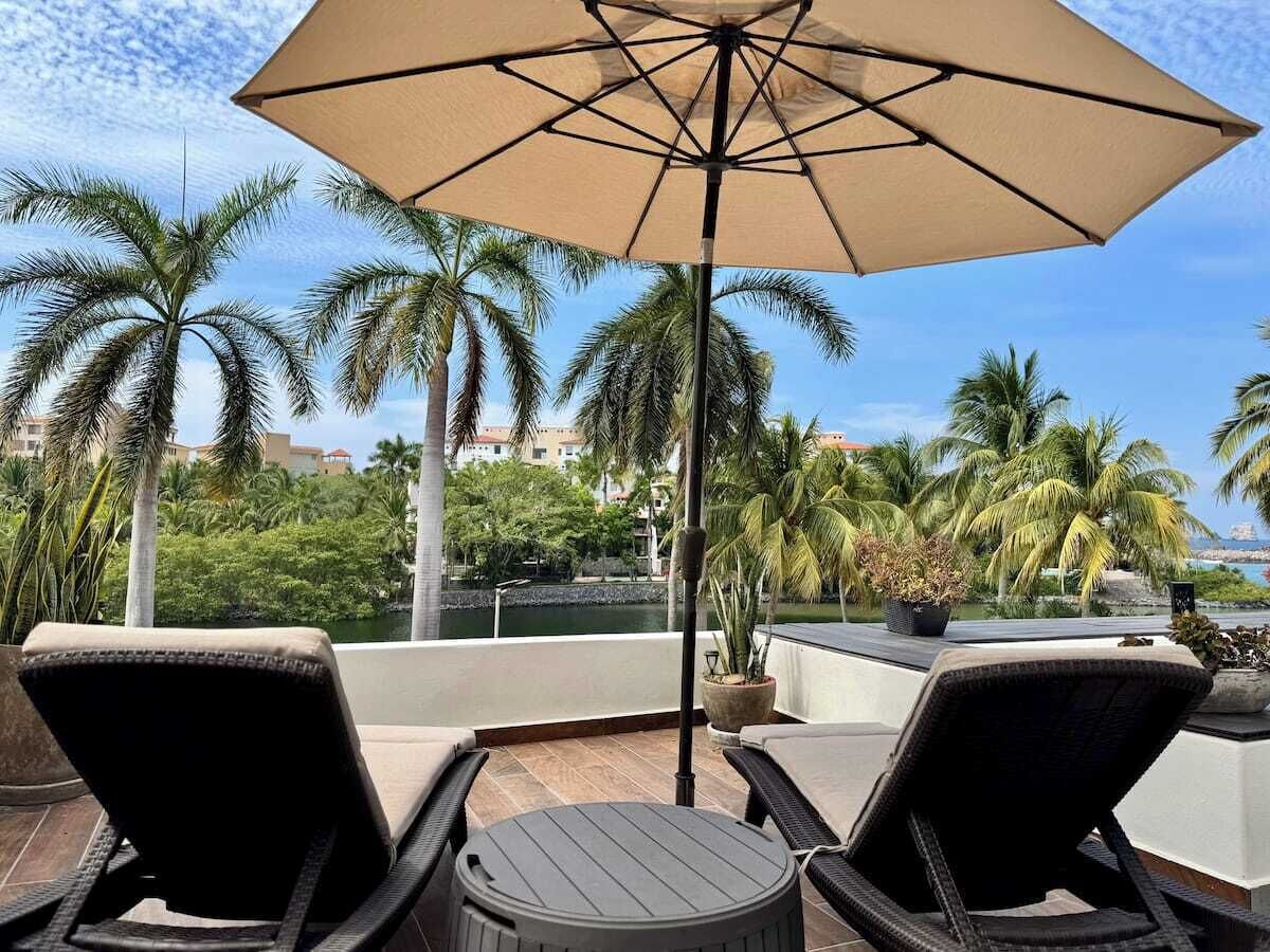 A terrace with two lounge chairs and a table, an umbrella, and a view of palm trees and buildings.