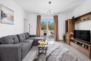 A modern living room with a grey sectional sofa, a round coffee table with fruits and snacks, a flat-screen TV on a wooden stand, and a large window with curtains. - Holiday House Lana With Pool, Biograd na Moru, Croatia (Biograd na Moru)