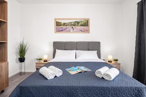 Ein ordentlich bezogenes Bett mit blauer, gesteppter Decke, zwei aufgerollte Handtücher, einer Reisemagazin und einem gerahmten Landschaftsbild darüber.