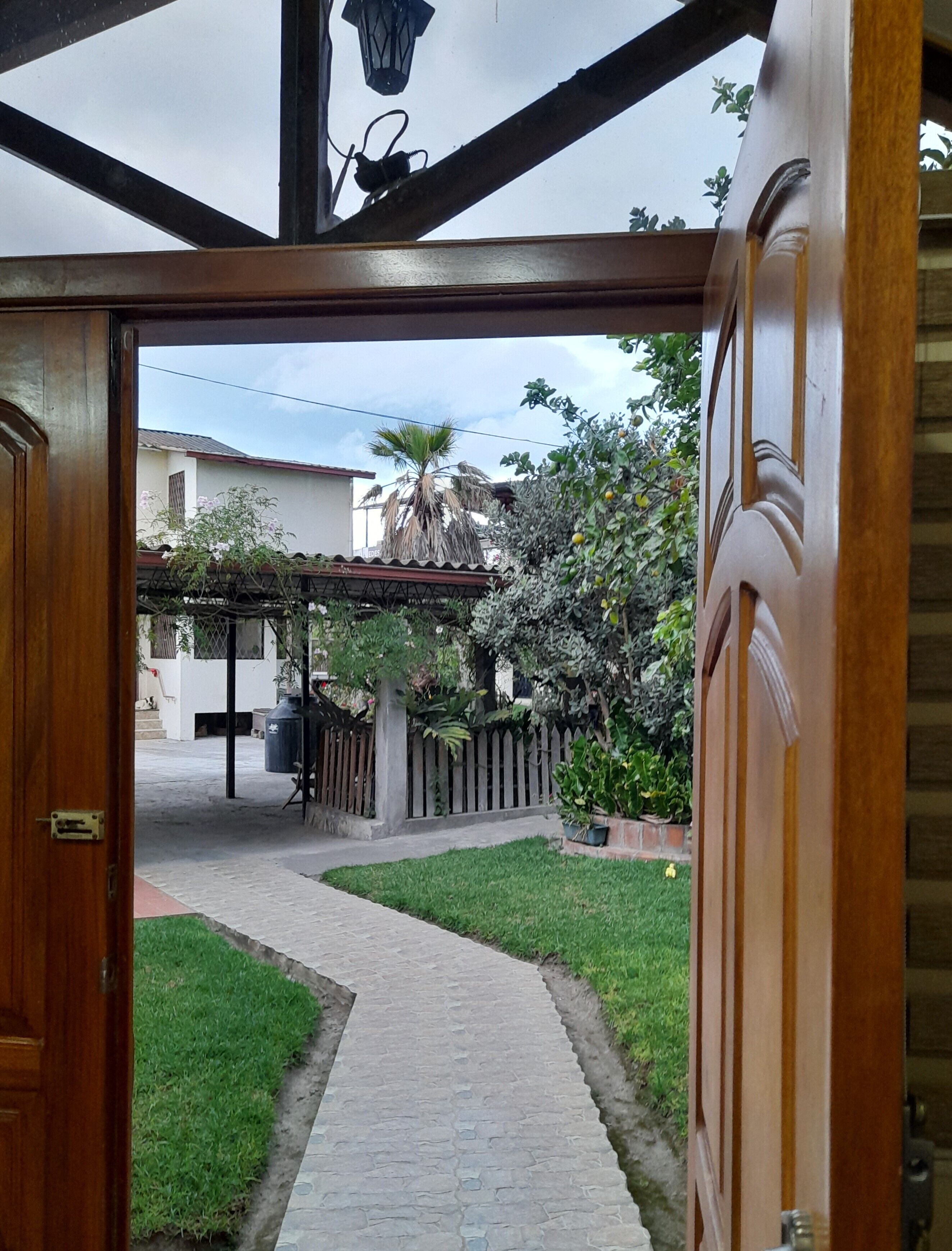 Una puerta de madera abierta que da a un camino empedrado, rodeado de follaje y una cerca de madera.